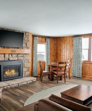 Cozy living space featuring a stone fireplace, wooden accents, a circular rug, a dining table, and curtains framing bright windows.