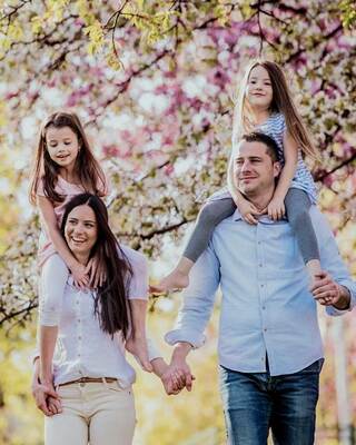 A joyful family walks together under blooming trees, with two girls on their parents' shoulders, enjoying a beautiful day.