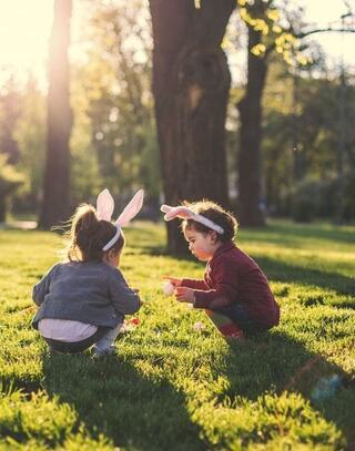 Two kids wearing bunny ears play together on grassy ground, basking in warm sunlight in a serene park setting.