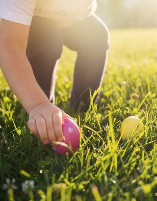 A child's hand reaches for colorful Easter eggs in a sunlit grassy field, capturing the joy of a playful egg hunt.