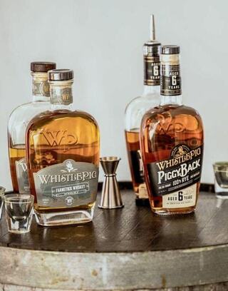Two bottles of WhistlePig whiskey and PiggyBack rye sit on a wooden barrel, accompanied by a jigger and shot glasses.