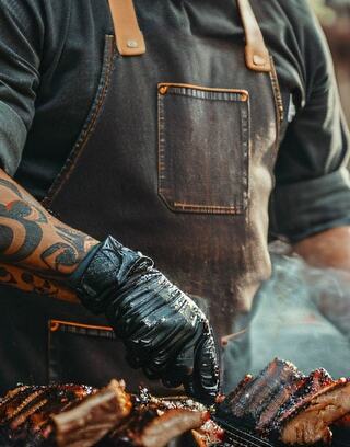 A tattooed chef in a dark apron and gloves skillfully handles smoky, grilled ribs on a barbecue.