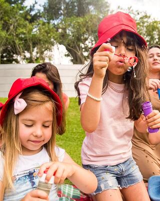 Two young girls in matching red hats play outdoors, one blowing bubbles while the other focuses on a bottle, surrounded by friends.