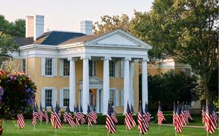 A stately yellow mansion with grand pillars and a manicured lawn, adorned with numerous American flags and lush green trees.