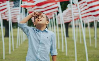 A young boy stands in a field of flags, saluting while looking upward, evoking a sense of pride and reflection.