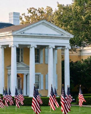 A large, elegant building with white columns stands behind a manicured lawn adorned with multiple American flags. Green trees provide a natural backdrop.