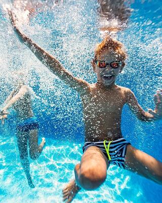 A young boy with sunglasses smiles and splashes underwater, surrounded by shimmering bubbles, while another child swims nearby.