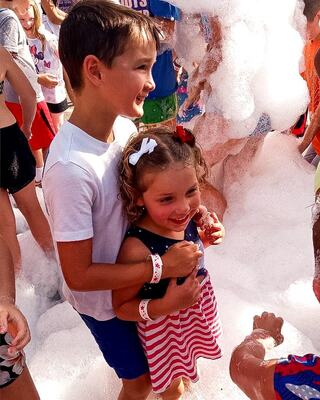 Two smiling children enjoy a playful foam party, surrounded by other kids in colorful outfits, creating a fun and cheerful atmosphere.
