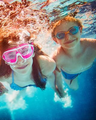 Two children swim underwater, smiling and wearing goggles, surrounded by glittering blue water. Joyful moments captured in a fun aquatic setting.