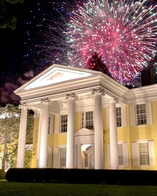 A grand yellow mansion with white columns shines brightly at night, illuminated by vibrant fireworks bursting in colorful splendor above.