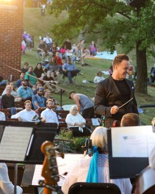 A conductor engages with an outdoor orchestra, while an audience enjoys the performance on a grassy hillside beneath a warm sunset.