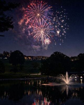 Colorful fireworks burst over a serene pond, reflecting vibrant lights, with a fountain adding to the enchanting night scene.