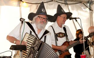 Two musicians perform in German festive attire, one with an accordion and the other with a guitar, in a lively tent setting at Oglebayfest.