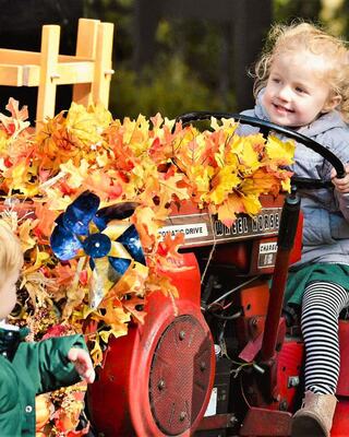 A joyful child drives a decorated tractor adorned with colorful autumn leaves, while another child plays nearby.