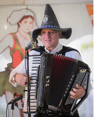A man in traditional attire plays the accordion on stage, wearing a large felt hat, with lively decorations in the background.