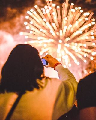 A person captures a vibrant fireworks display on their phone, surrounded by a crowd enjoying the festive atmosphere.