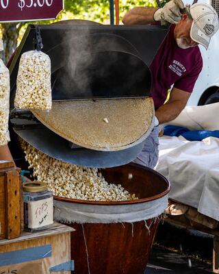 A vendor pours freshly made popcorn from a large machine into a wooden bowl, creating a cloud of steam in a lively market setting.
