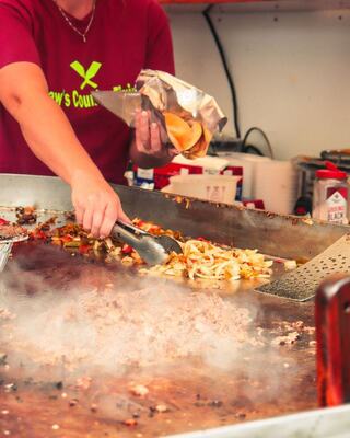 A person with a tongs is cooking on a large griddle, mixing meat, vegetables, and tortillas, creating a flavorful street food scene.