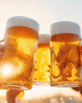 Three frosty mugs of beer are raised cheerfully, glimmering in the sunlight, creating a celebratory atmosphere.