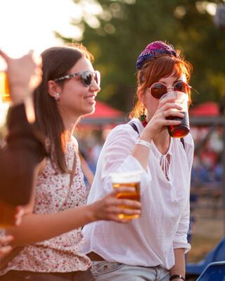 Two women enjoy drinks at a lively outdoor setting, wearing stylish sunglasses and engaging in conversation as the sun sets.