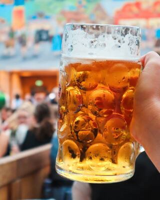 A hand holds a frosty beer mug, with a lively crowd and colorful mural in the background, capturing a festive atmosphere.