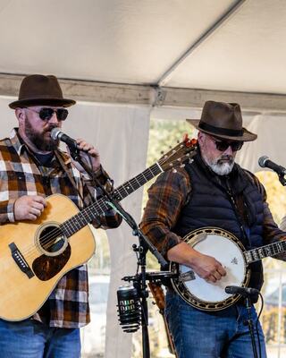 Two musicians in hats perform on stage, one with an acoustic guitar and the other with a banjo, both dressed in plaid shirts.