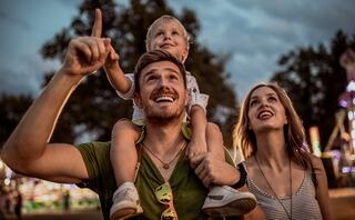 A happy family enjoys a night at the fair, with a father pointing excitedly and a child on his shoulders, while the mother smiles nearby.