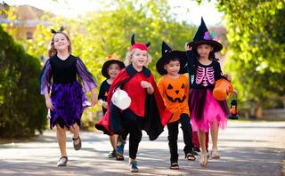 A group of children joyfully dressed in Halloween costumes, including witches, a skeleton, and a pumpkin, are walking along a sunny path.