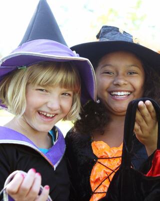 Two young girls in witch costumes laugh joyfully, one in a purple hat and the other in orange, sharing a fun Halloween moment together.