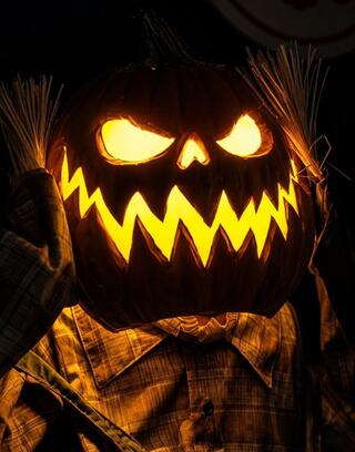 A glowing, menacing jack-o'-lantern with sharp teeth and fierce eyes, held by a figure wearing a plaid shirt and straw hair. Decorations that can be seen at Oglebay Good Zoo's Boo at the Zoo event.