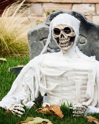 A skeleton dressed in white rags sits on the grass near a gravestone, creating a spooky Halloween atmosphere.