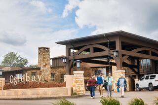A group of four people walks towards a rustic entrance featuring stone and wood architecture, with a sign reading "Oglebay."