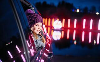 A smiling girl in a purple hat leans out of a car window, with colorful lights reflecting on a dark, tranquil lake in the background.