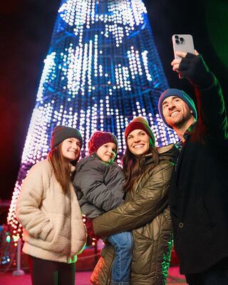 A joyful family takes a selfie in front of a brightly lit holiday tree, wearing winter hats and cozy jackets, embracing the festive spirit.