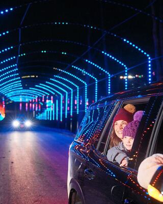 Two smiling faces peek out of a car window, surrounded by vibrant blue and colorful lights in a festive nighttime tunnel.