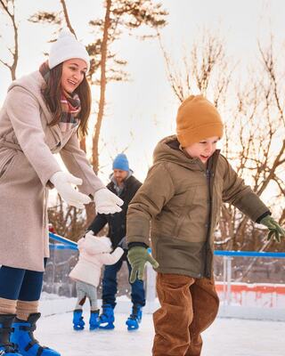 A woman encourages a young boy on skates, while another child and a man enjoy the winter activity, surrounded by trees in a snowy setting.
