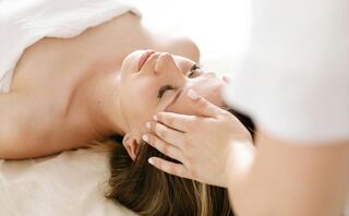 A woman relaxes during a facial massage, eyes closed and serene, as a therapist gently works on her head.