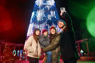 A cheerful family poses for a selfie in front of a brightly lit holiday tree, surrounded by festive decorations and glowing lights.