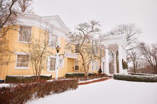 A cheerful group of four stands in front of a beautifully lit, festive mansion, surrounded by snow and holiday decorations.
