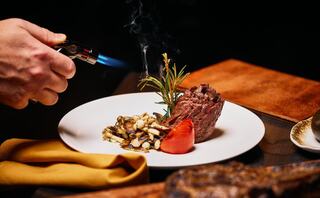 A hand uses a torch to caramelize a beautifully plated dish featuring steak, mushrooms, and a roasted tomato, garnished with fresh herbs.