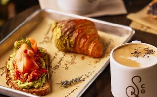 A tray features avocado toast topped with salmon and a croissant alongside a cup of latte decorated with lavender.