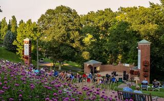 A scenic outdoor venue hosts a gathering, with people seated on a lawn, enjoying a performance surrounded by lush trees and blooming flowers.