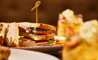 A stacked sandwich filled with layers of lettuce, tomato, and other ingredients, garnished with a skewer, accompanied by drinks.