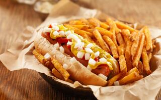 A hot dog topped with mustard, ketchup, and diced onions, served alongside crispy golden fries on parchment paper in a rustic bowl.