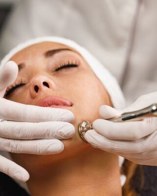 A woman receives a facial treatment, eyes closed, as a professional applies a tool for skin care with gloved hands.