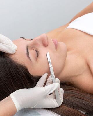 A woman receives a facial treatment, lying down with closed eyes, while a gloved hand uses a scalpel for skincare.
