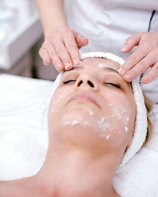 A person enjoys a facial treatment, with a professional applying a product while they recline on a treatment table, eyes closed in relaxation.