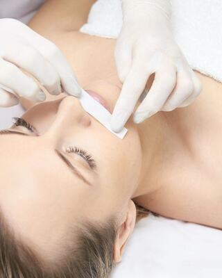 A woman is receiving hair removal treatment, with a gloved technician applying a wax strip to her upper lip.