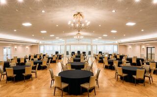 A spacious banquet hall with round tables covered in black cloth, surrounded by elegant chairs and bright lighting from a central chandelier.