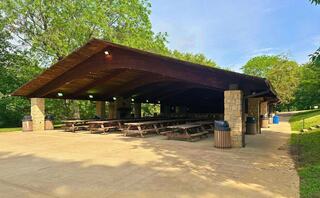 A spacious picnic shelter with a wooden roof and stone pillars. It features long picnic tables and trash bins, surrounded by greenery.